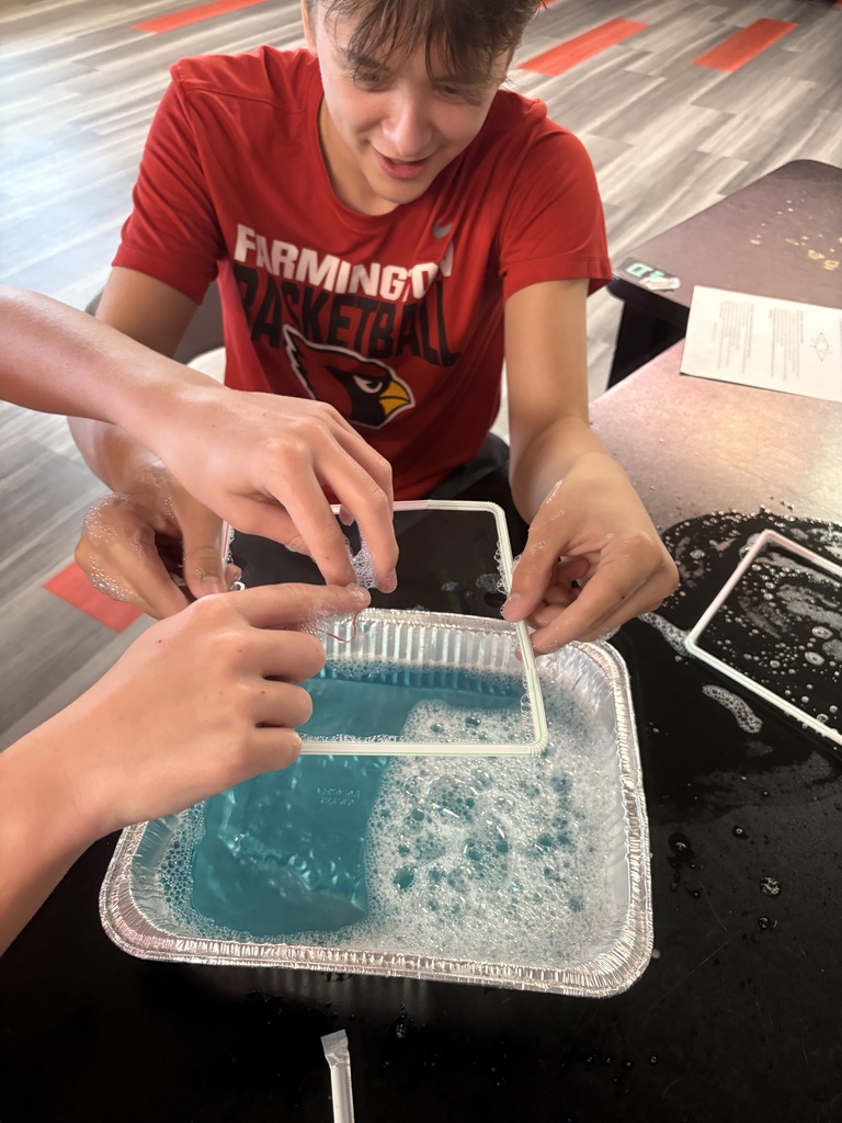 A person in a red t-shirt sits at a table with another person, both handling a tray with blue liquid.