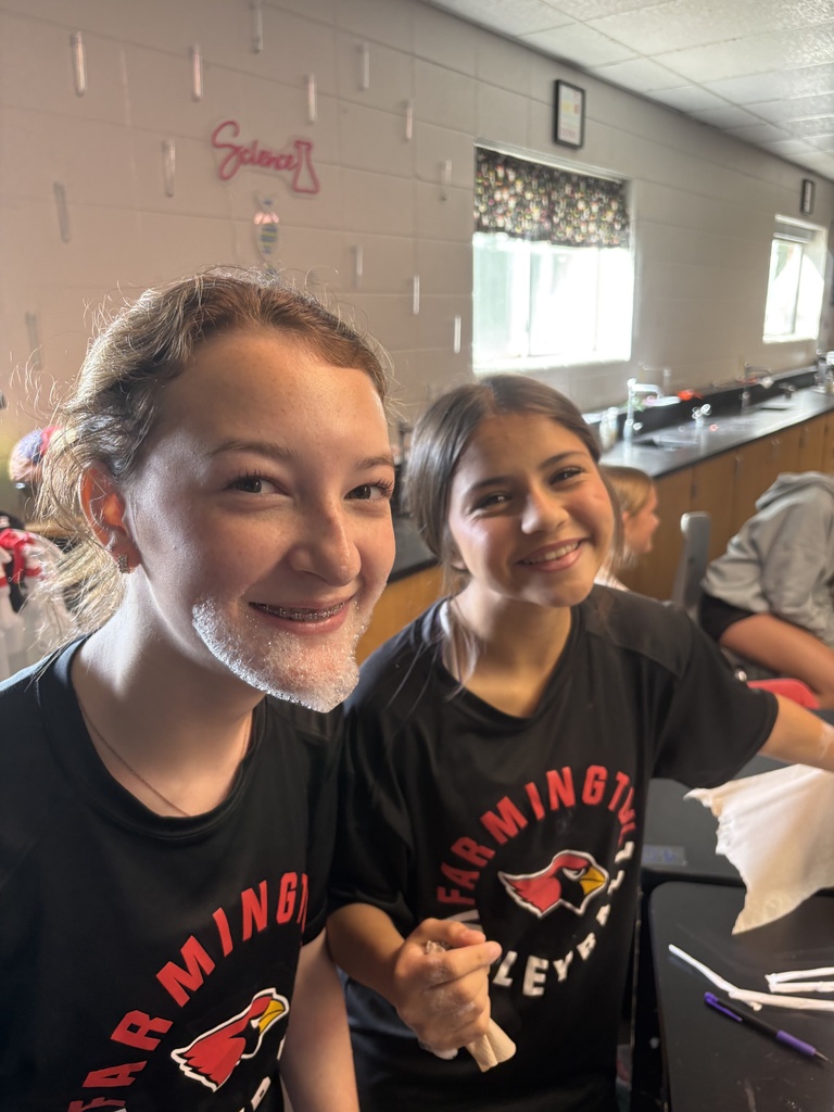 Two young women in black shirts with red and white emblems sit side by side, smiling. They appear to be in a classroom.