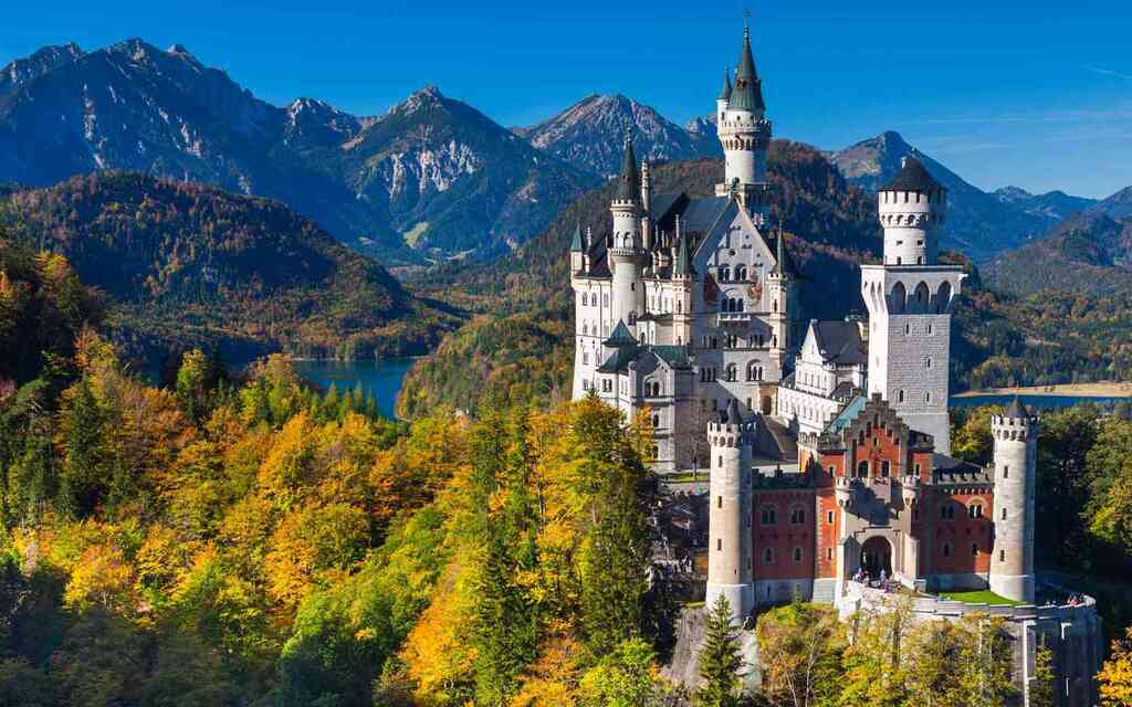 A castle with multiple towers and spires, set amidst mountains and surrounded by autumn trees.