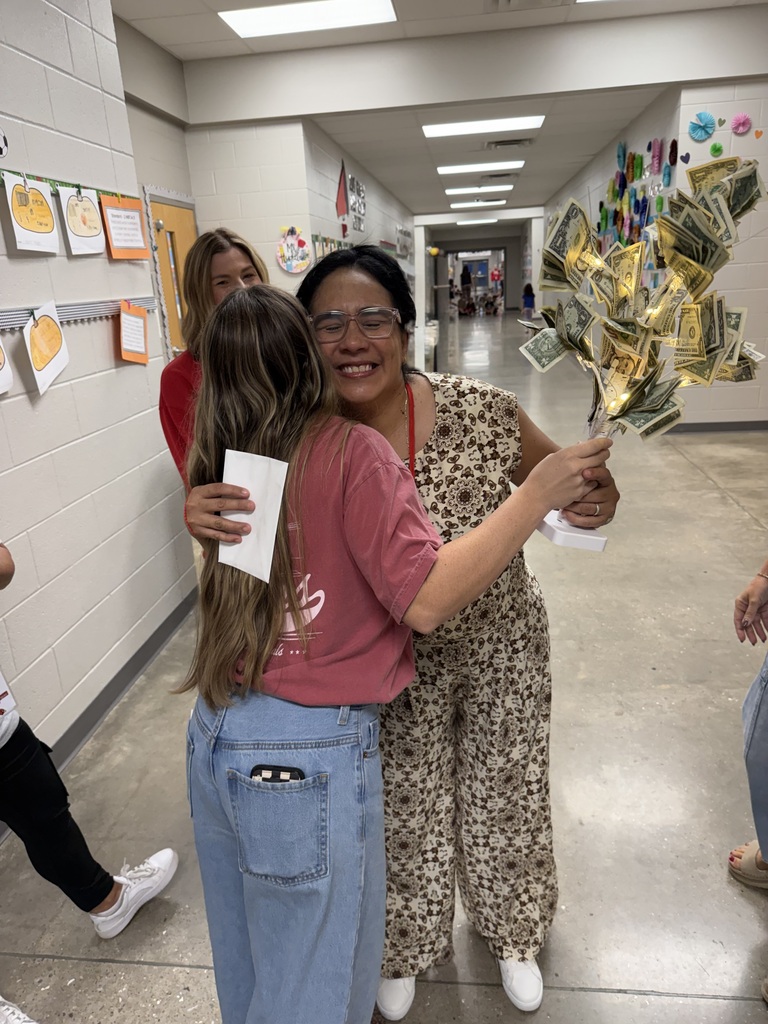 A woman holds money in a bouquet while hugging a girl who holds a paper in a school hallway.