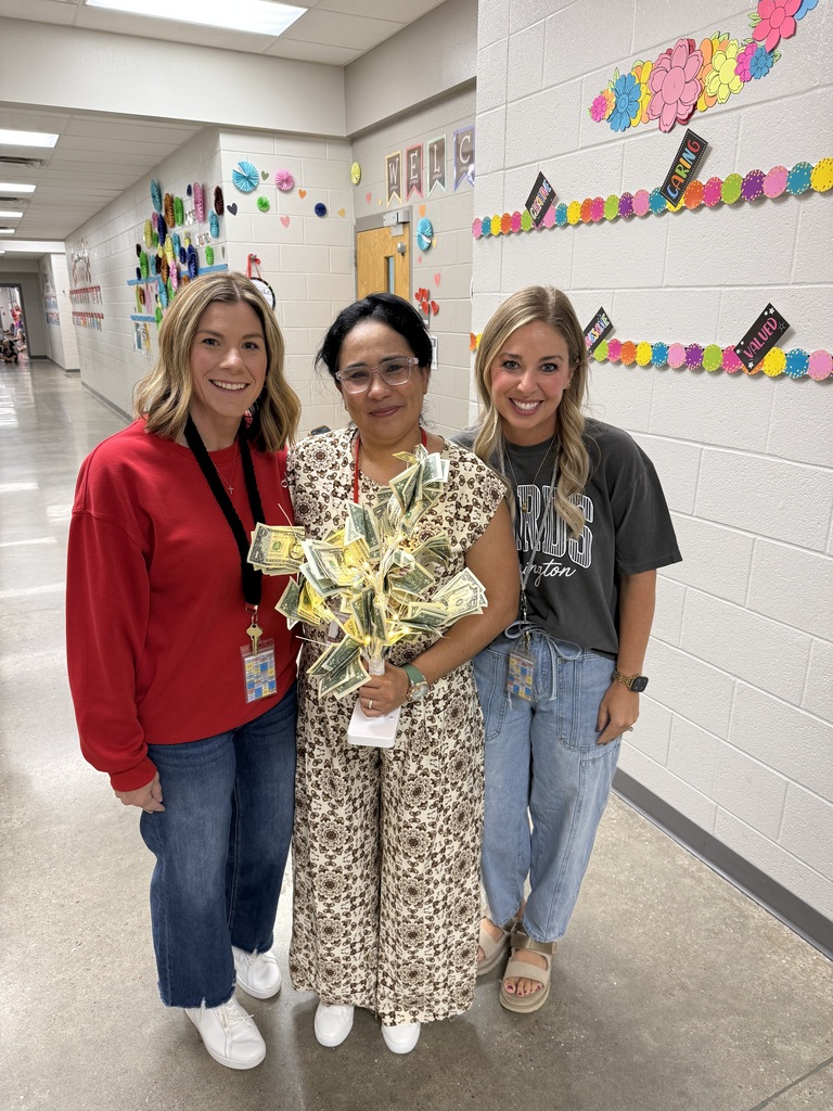Three women stand in a school hallway, smiling. One holds a gift. White walls with colorful decorations.