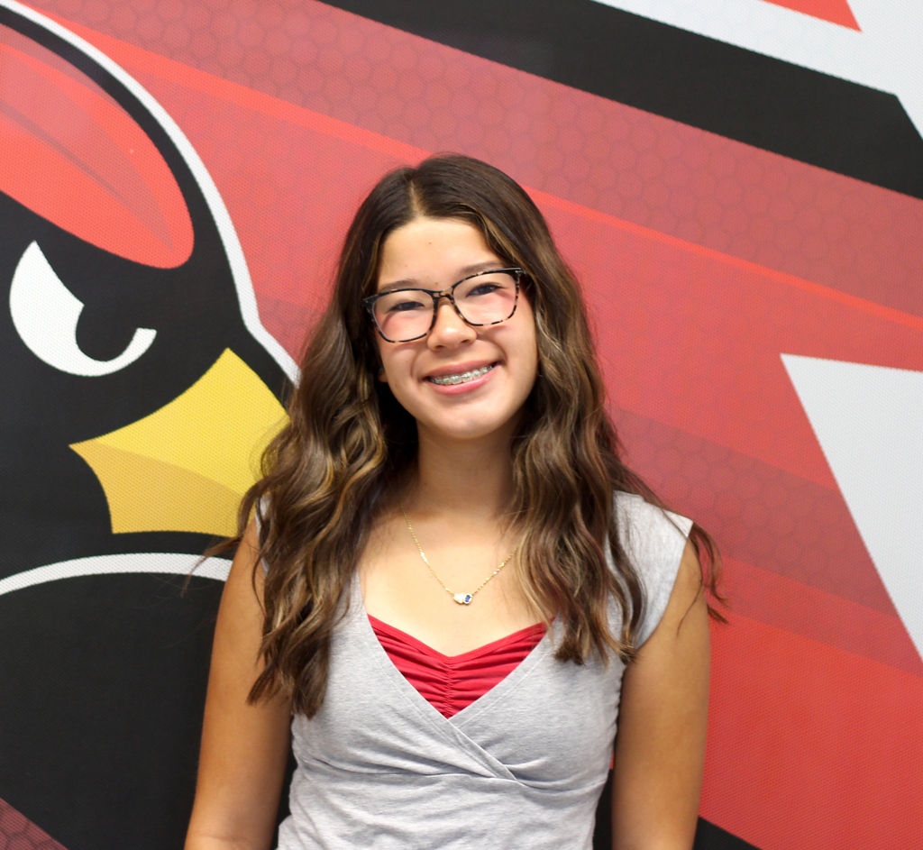 Woman with long hair wearing glasses and a necklace stands before a red and black wall with a logo.