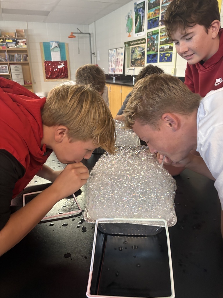 Four children, two in red, one in white, and one in black, focus on a plastic bag filled with ice.
