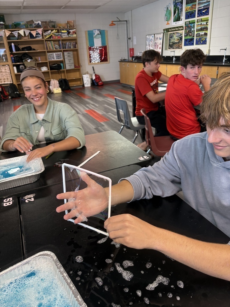 Students sit at tables with various items; one holds a clear plastic cube, another has a tray with blue liquid.