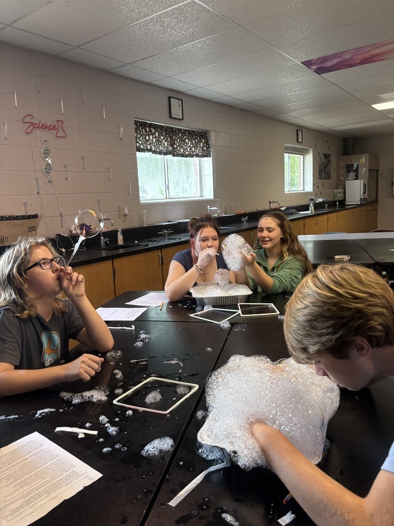 Four students sit at a table in a classroom. Three of them blow bubbles from a bubble maker.