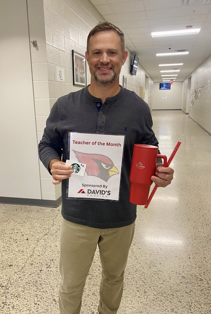 A man in a hallway holds a "Teacher of the Month" sign and a red cup. He wears a black long-sleeve shirt, khaki pants, and a smile.