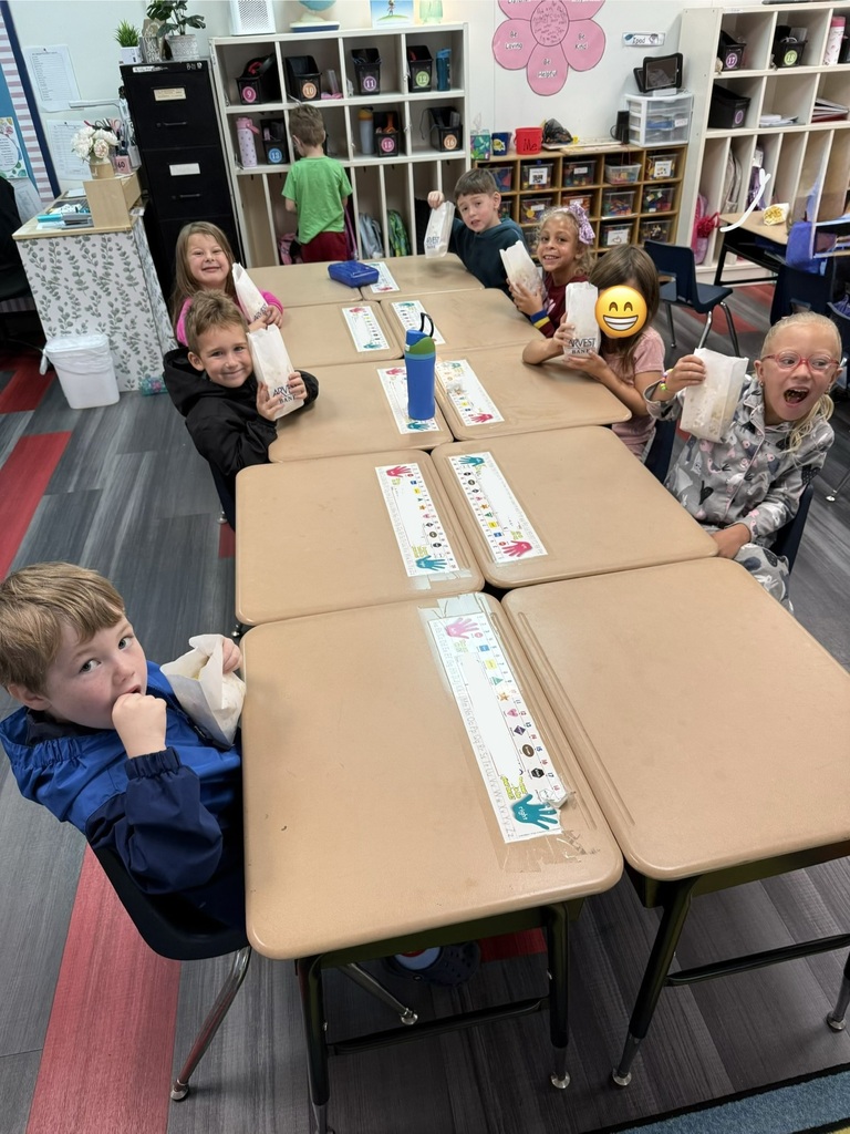 A classroom with children seated around desks, smiling, and holding items. Background features shelves, tables, and a red floor.