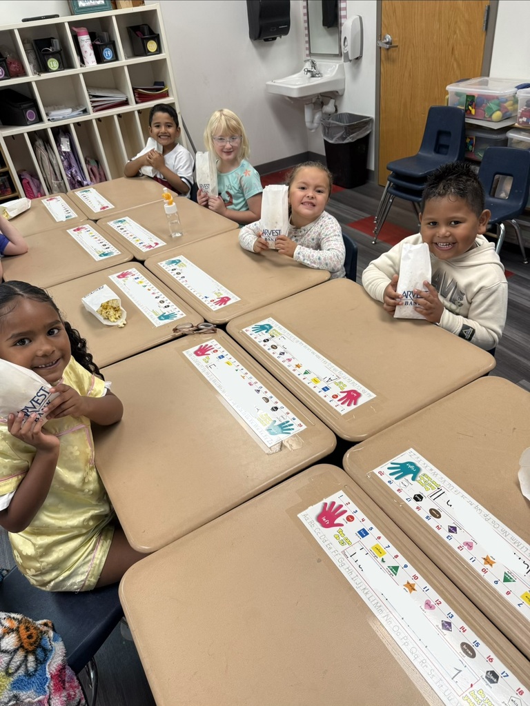 Five children in a classroom setting, seated at desks, smiling and holding food items. Desks have printed papers with handprints.