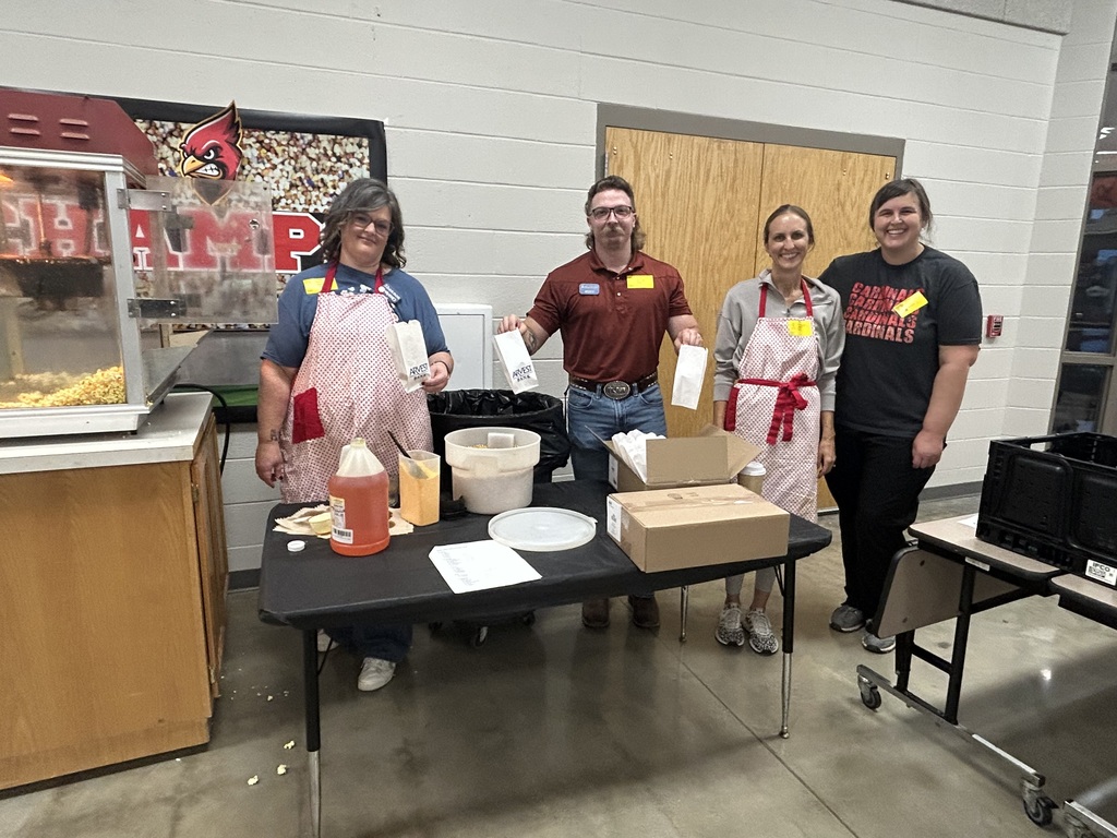 Four people in aprons stand around a table with boxes, food, and containers. A popcorn machine and a wall with posters are nearby.