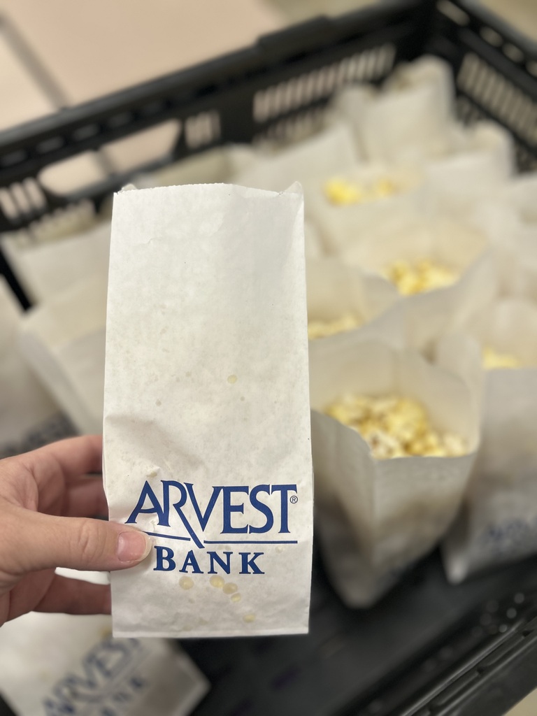 A hand holds a bag of popcorn. The bag says "ARVEST BANK". Many bags are stacked in a black crate.