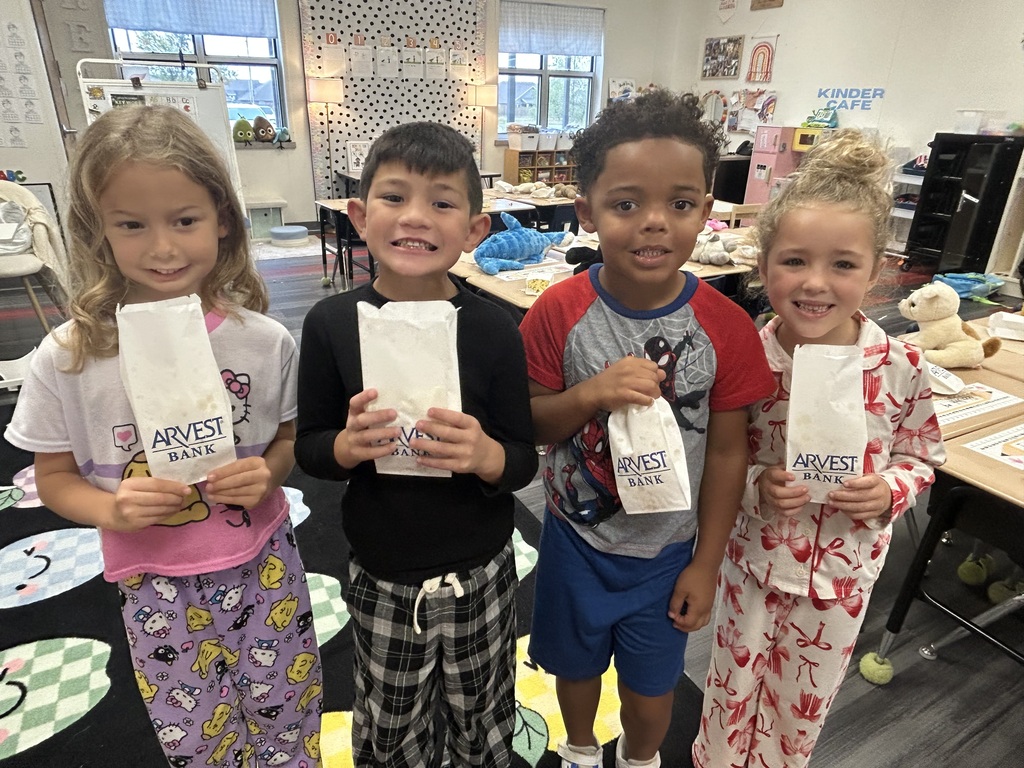 Four children in pajamas, two girls and two boys, stand together holding bags. Behind them, desks and chairs are set up, and a window lets in natural light.