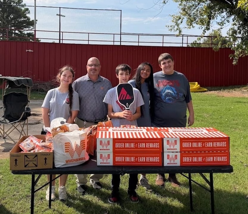 A group of people pose for a photo in front of a table with boxes. A grassy field and red fence are behind them.