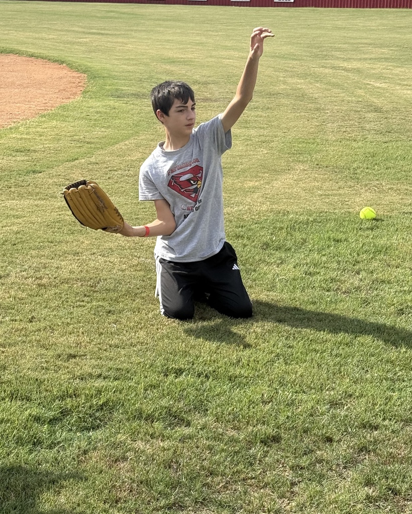 A person kneels on grass, holding a baseball glove, and throws a ball in an outdoor sports field.