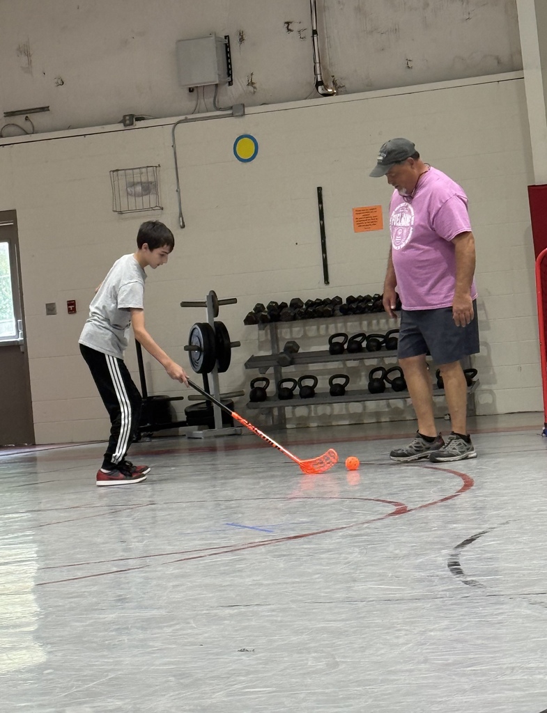 A boy plays with a hockey stick on an indoor court. An adult stands behind him, wearing a hat and sneakers.
