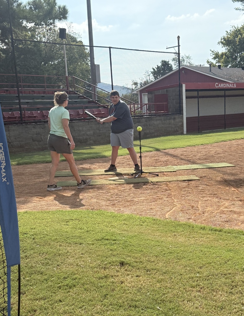 Two people practice hitting a baseball at a practice field, with a red building in the background.