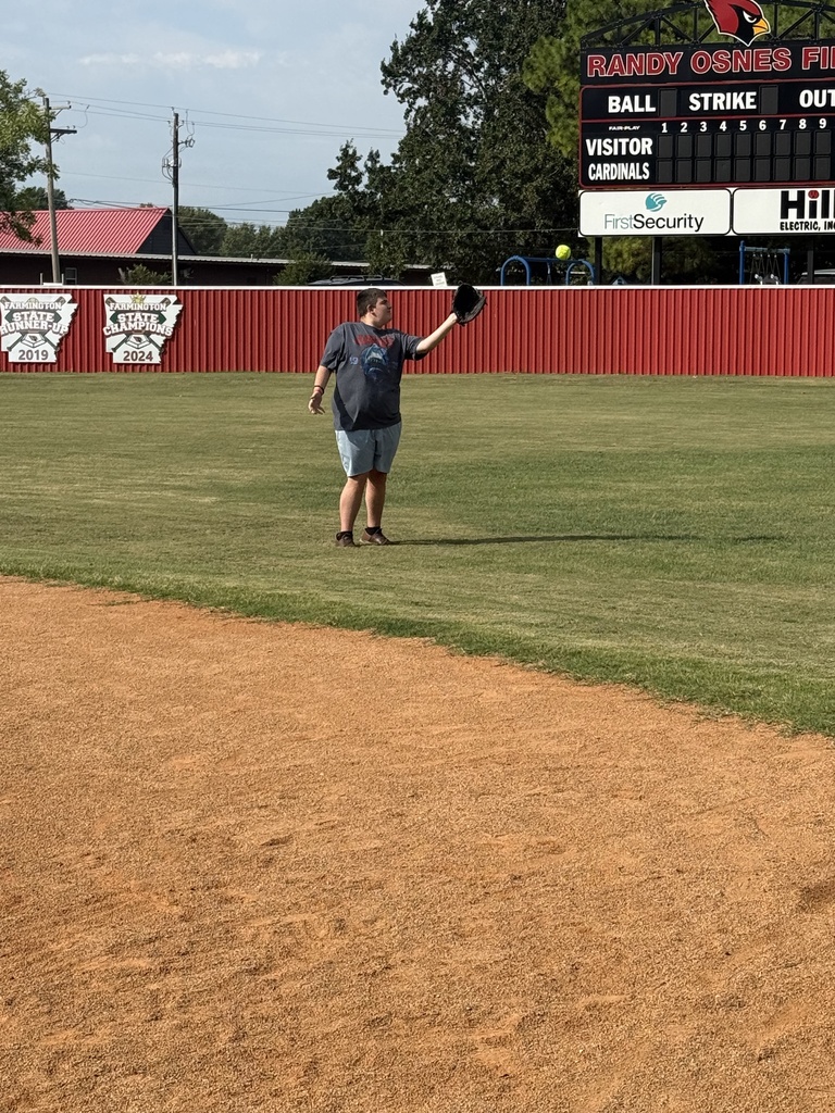 A person stands on a baseball field with a baseball glove, near a red wall with a scoreboard.