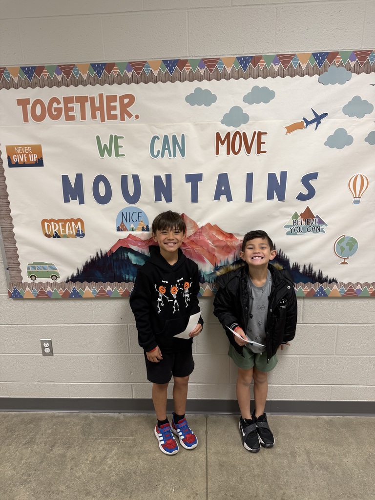 Two young boys stand together, smiling and holding items, in front of a wall banner with mountains, clouds, and balloons.