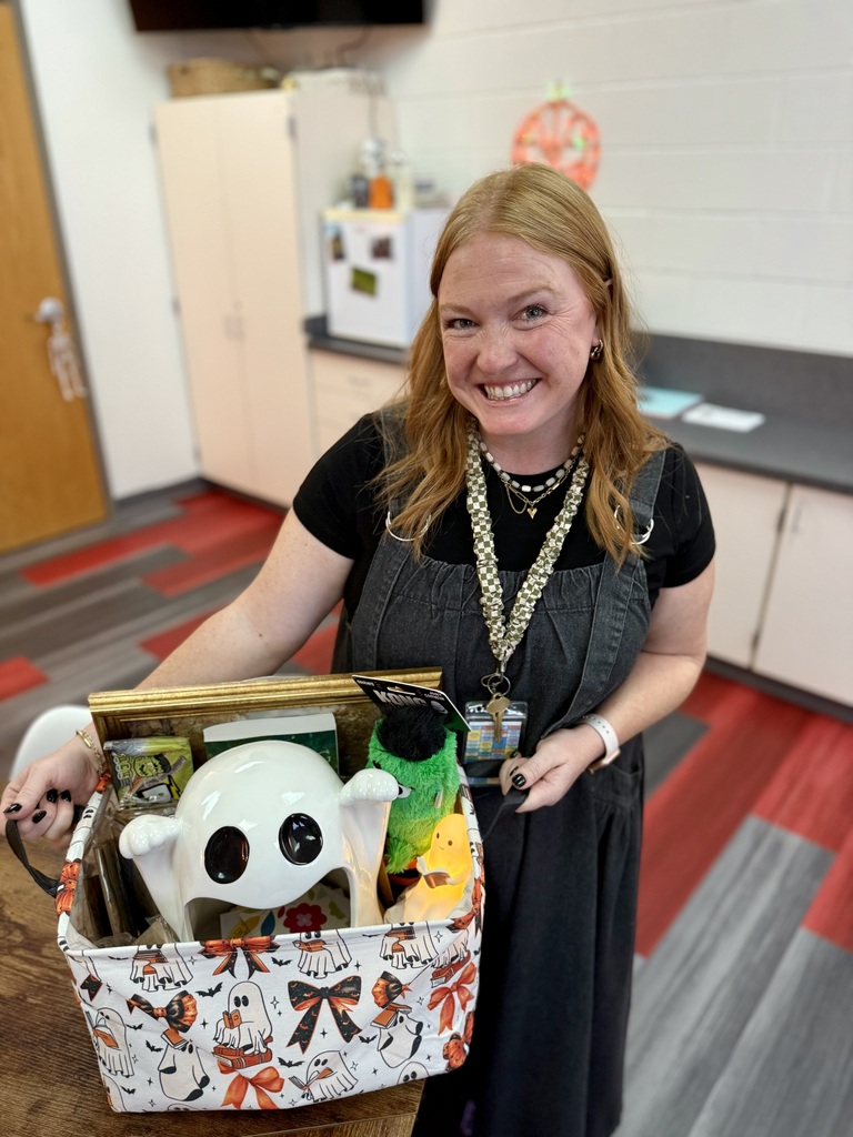 A woman holds a basket with various toys and a pumpkin. She stands in a kitchen with a refrigerator, cabinets, and red floor tiles.