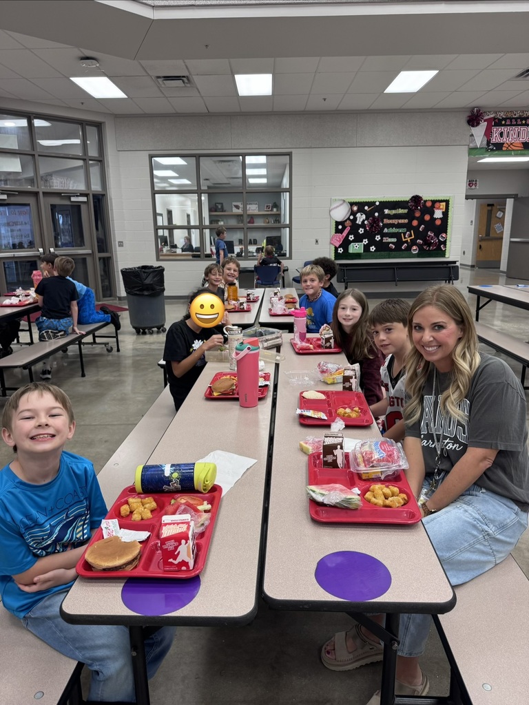 A group of children in a cafeteria. Some have trays of food. A teacher sits at the table.
