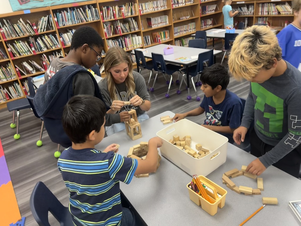 A group of children are building with blocks at a table in a library with bookshelves in the background.