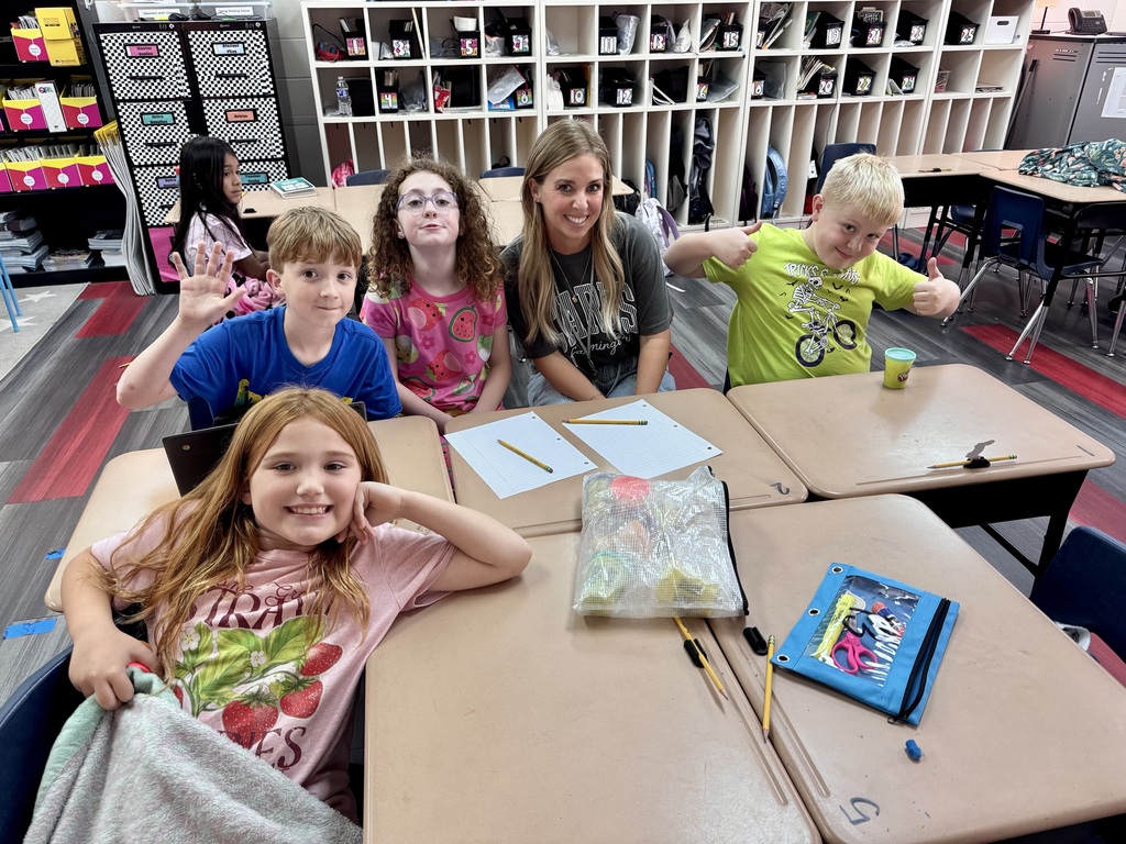 A classroom with children sitting at desks, smiling, and gesturing. One desk has a tablet, a bag, and a cup.