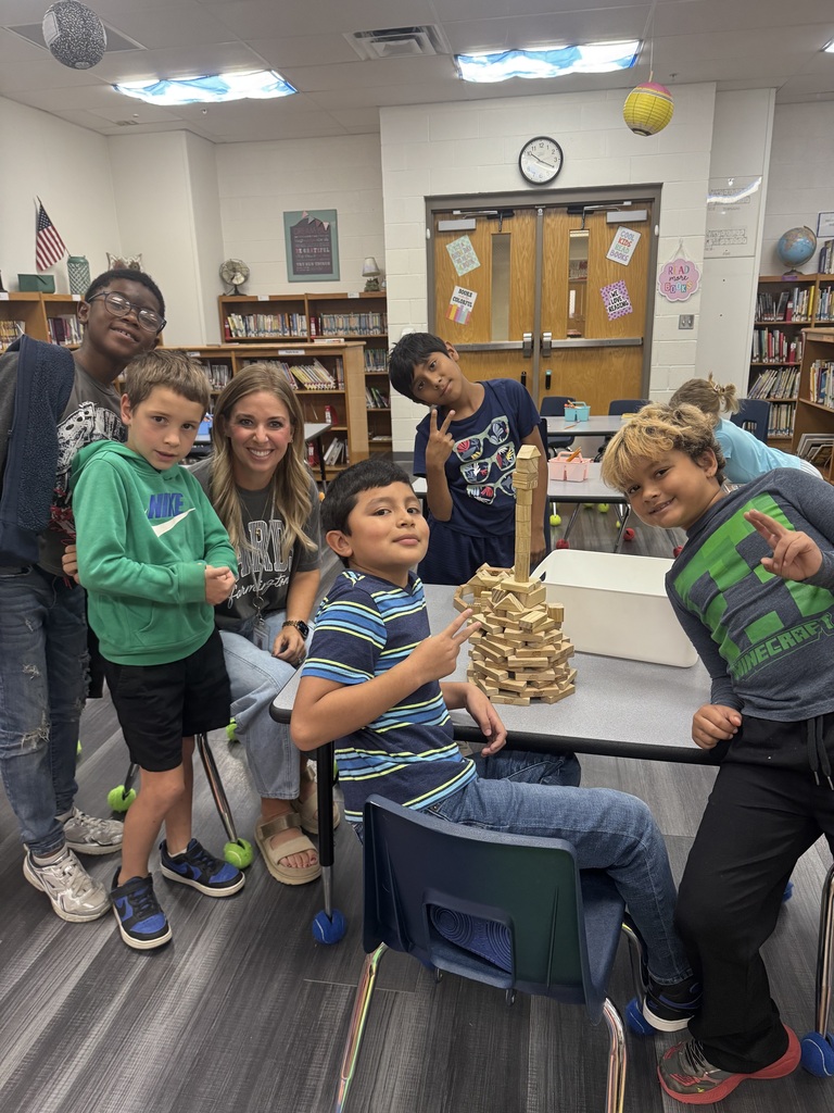Students are in a library, standing and sitting around a table with a stack of blocks.