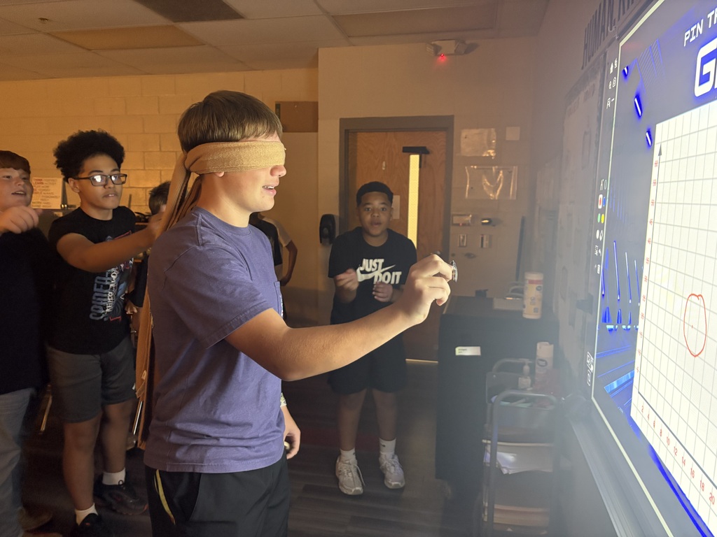 A young man with a blindfold stands at a whiteboard with a chart. Others watch as he points to the board.