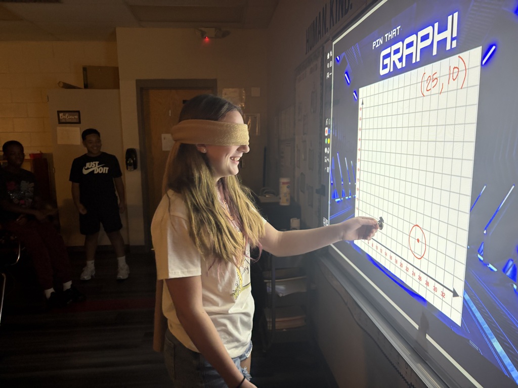 A person wearing a blindfold writes on a whiteboard with a chart labeled Graph. Another person stands behind them.