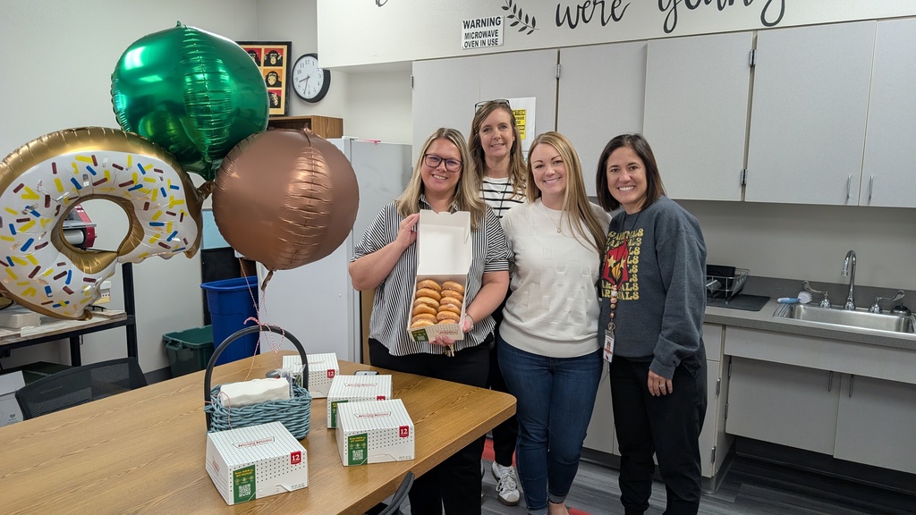 Four women pose with a table of boxes and balloons in a kitchen. A donut-shaped balloon is behind them.