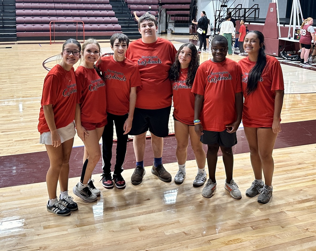 Seven individuals wearing red shirts and sneakers stand on a basketball court with a wooden floor.