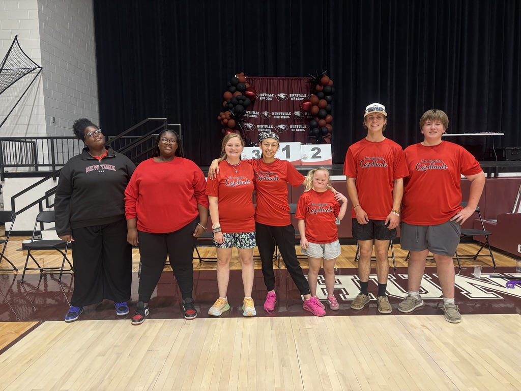 Group of people wearing red shirts and sneakers, standing on a basketball court, with a dark stage backdrop.