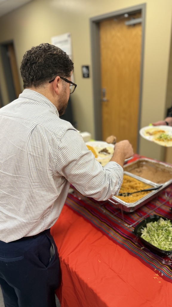 A man stands at a buffet table with a red tablecloth, wearing glasses and a striped shirt.