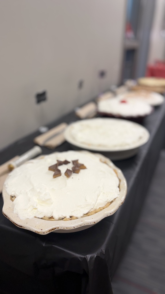 Long table with various pies on it, including chocolate-covered ones. White tablecloth and white walls in the background.