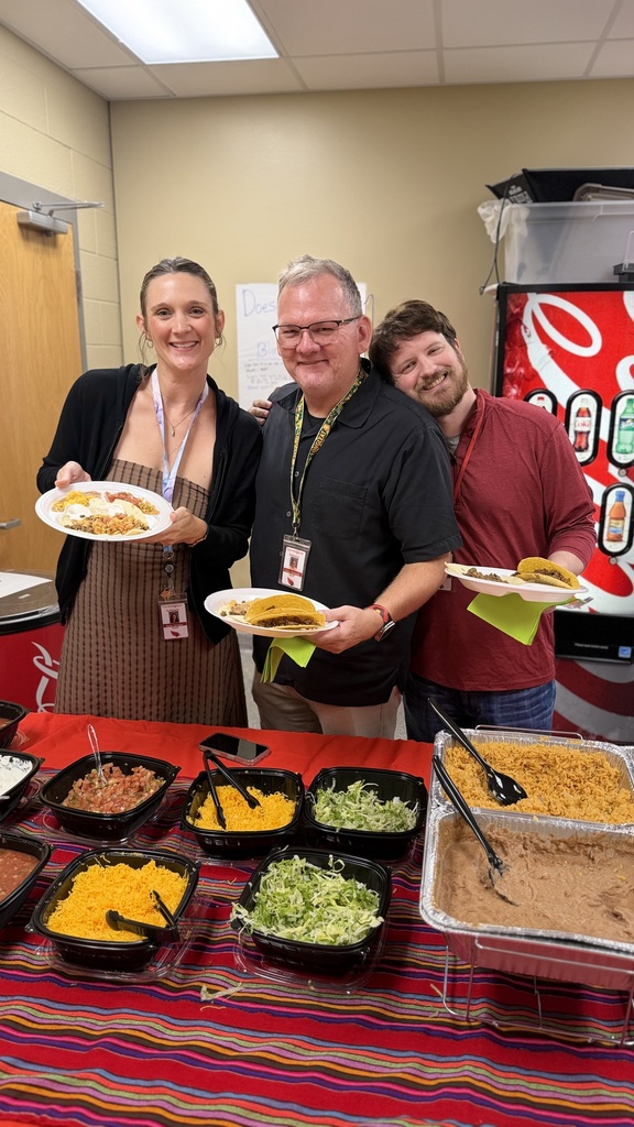 Three people smile as they hold plates of food at a buffet table with multiple dishes.