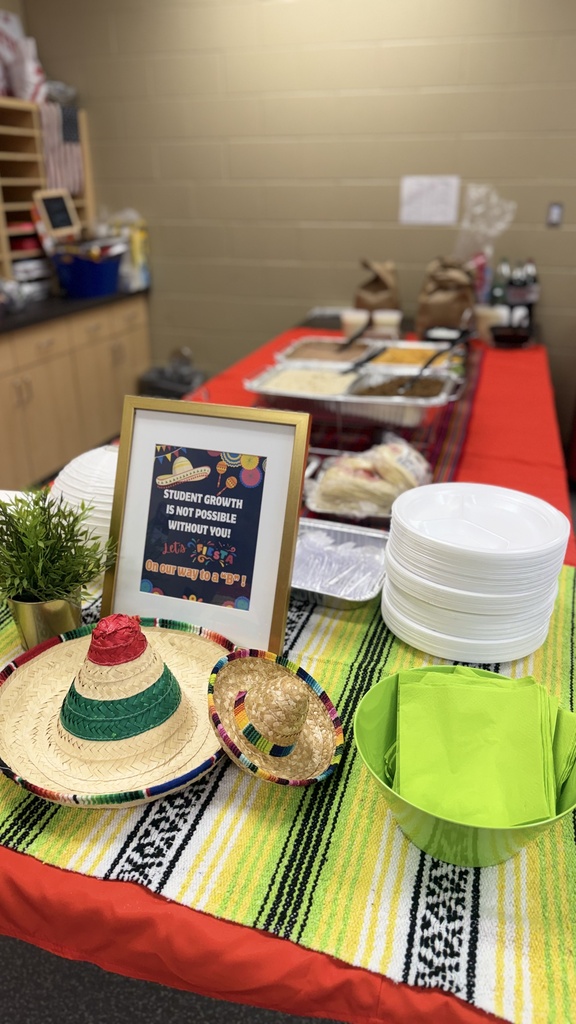 A table displays Mexican food with plates, a frame, sombreros, and a green bowl. Behind it, a wall shows a sign and shelves.