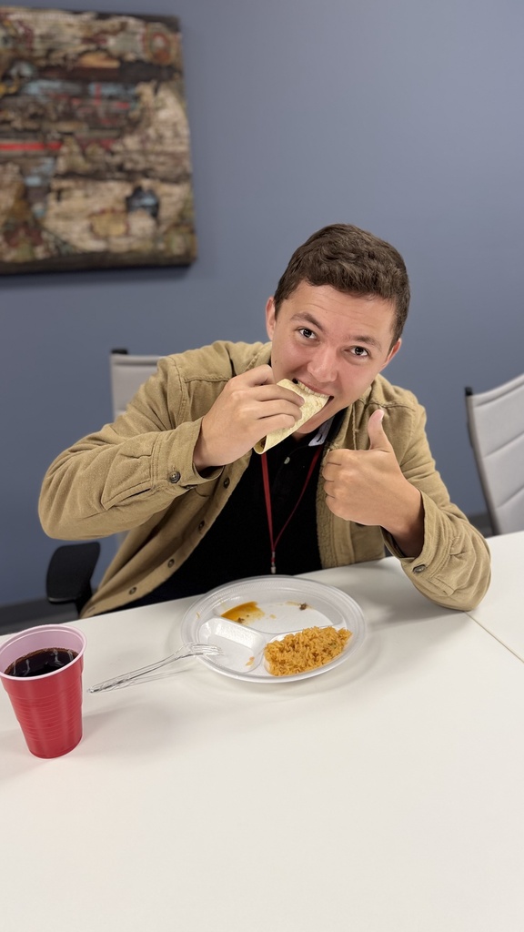 A person sitting at a table eating a sandwich and giving a thumbs up with food, a drink.
