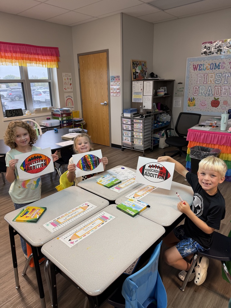 Kids sit at desks, displaying colorful posters with drawings. The classroom has desks, chairs, and a bulletin board.
