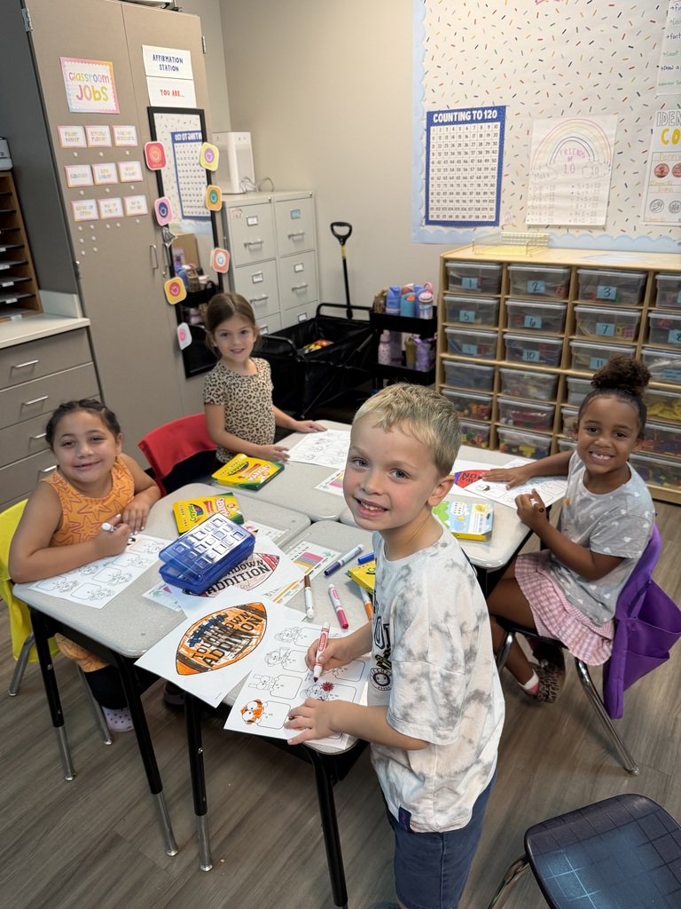 Four children are seated at tables in a classroom, working on coloring activities. A table holds coloring supplies.