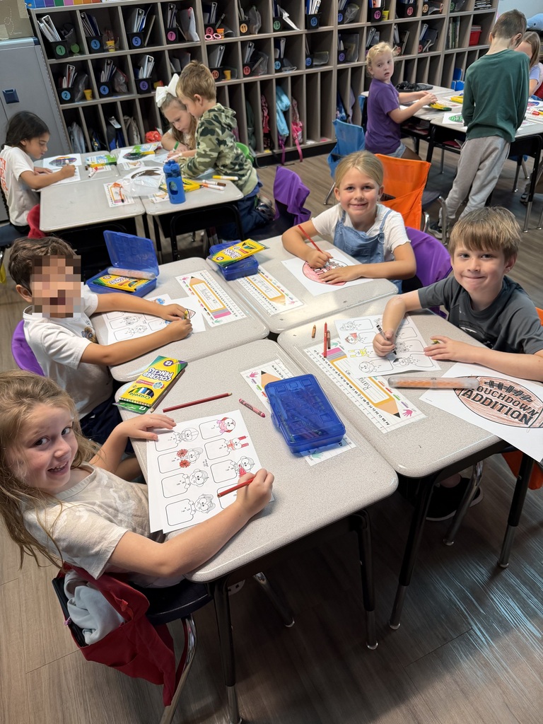 Classroom of students seated at desks, engaging in activities with art supplies. Shelves with books and materials in the background.