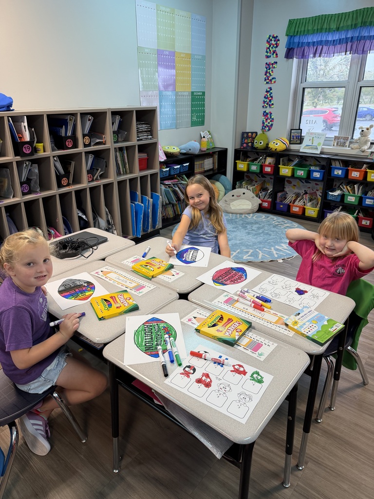 Three children are seated at a table with art supplies. A bookshelf, colorful papers on walls, and a window are in the background.