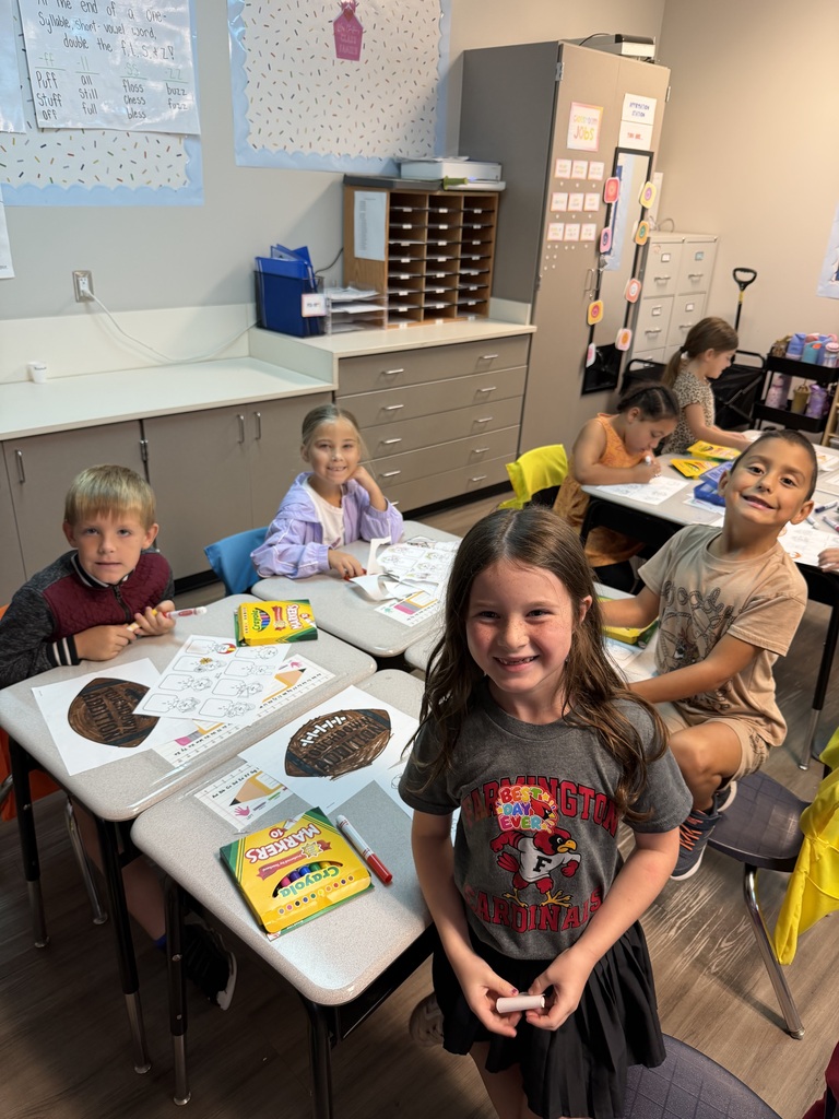 Children seated at desks, with one standing and smiling, working on art projects in a classroom.