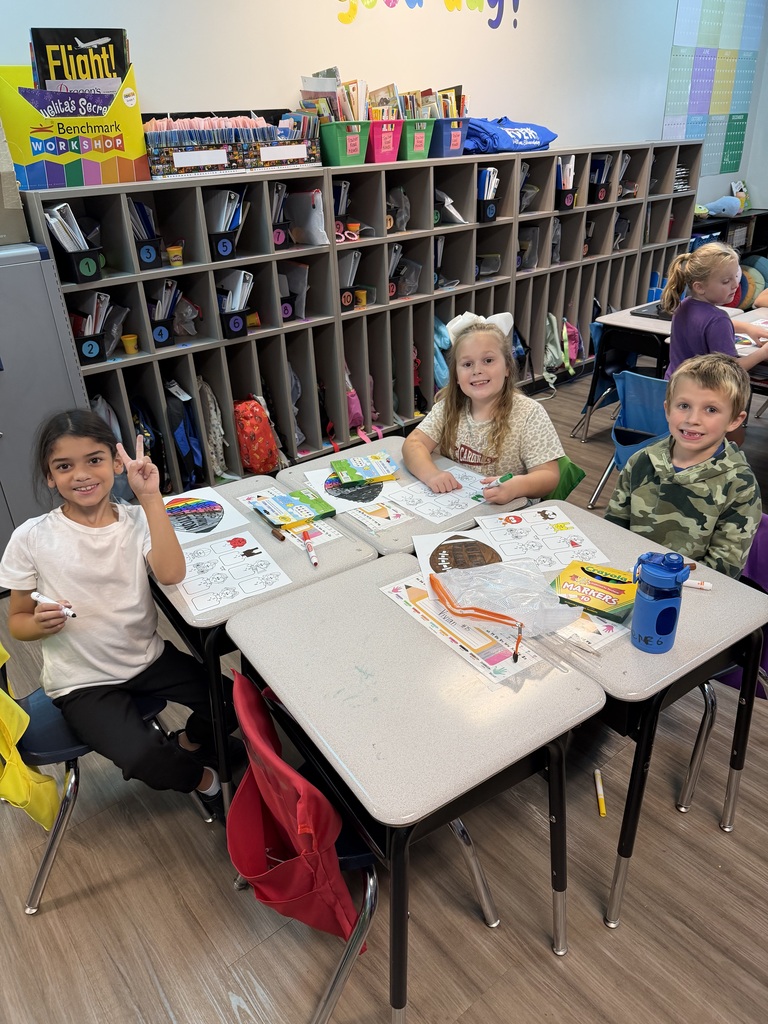 Three children sit at desks in a classroom. They have various supplies on the table. Behind them, a shelf holds books and supplies. A motivational sign is on the wall.