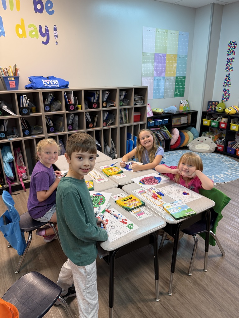 Four children sit at a table in a classroom with a teacher. Each child has a craft project.