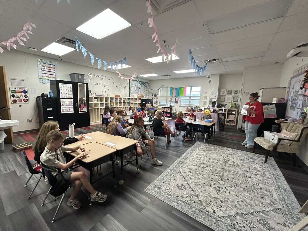 A classroom with students seated at tables. A teacher stands near a whiteboard. Rugs and chairs are on the floor.