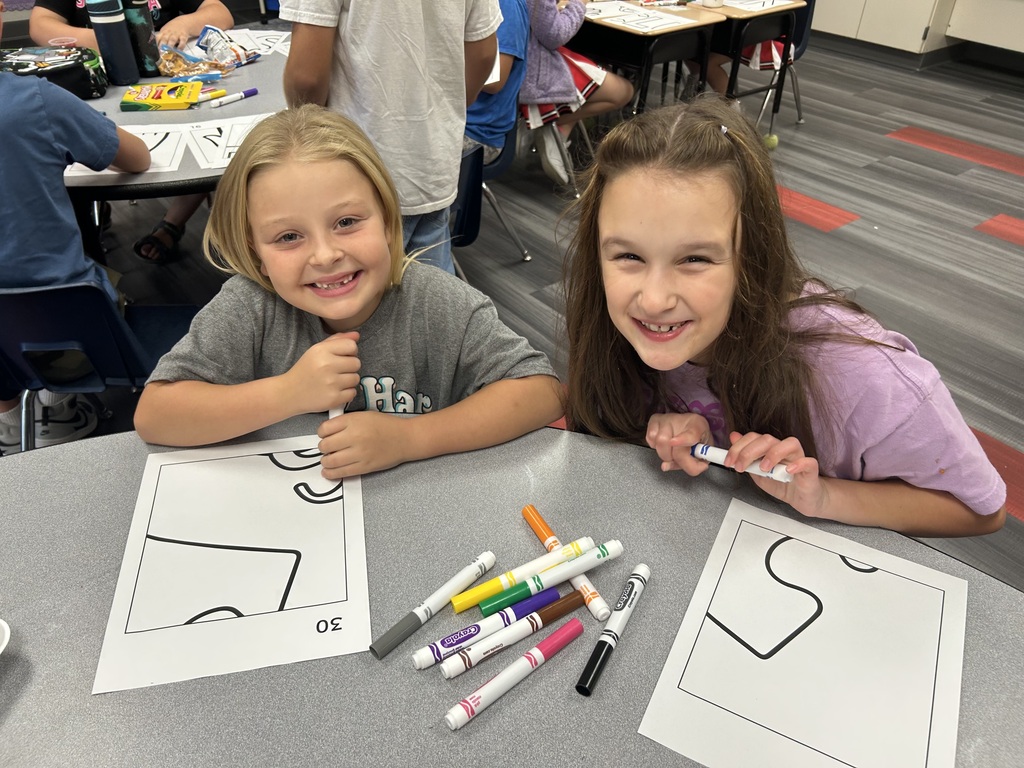 Two girls, one in gray and one in purple, smile and sit at a table with coloring books.