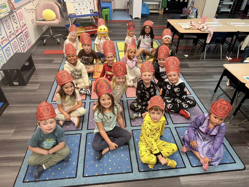 Children sitting in rows on a rug with red hats, some in pajamas, in a classroom with desks.