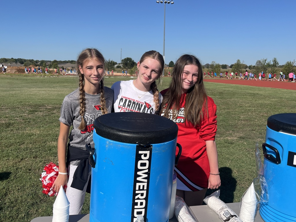 Three young women stand behind a blue barrel labeled "POWERADE" on a grassy field. They hold pom-poms and wear school spirit colors.