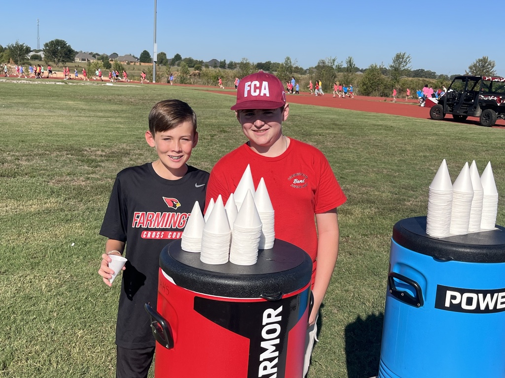 Two children stand with a container of cups, one holding a cup, on a sports field.