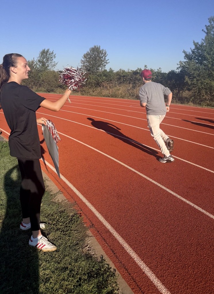 A woman with pom-poms stands at the side of a red track. A person runs in the opposite direction.