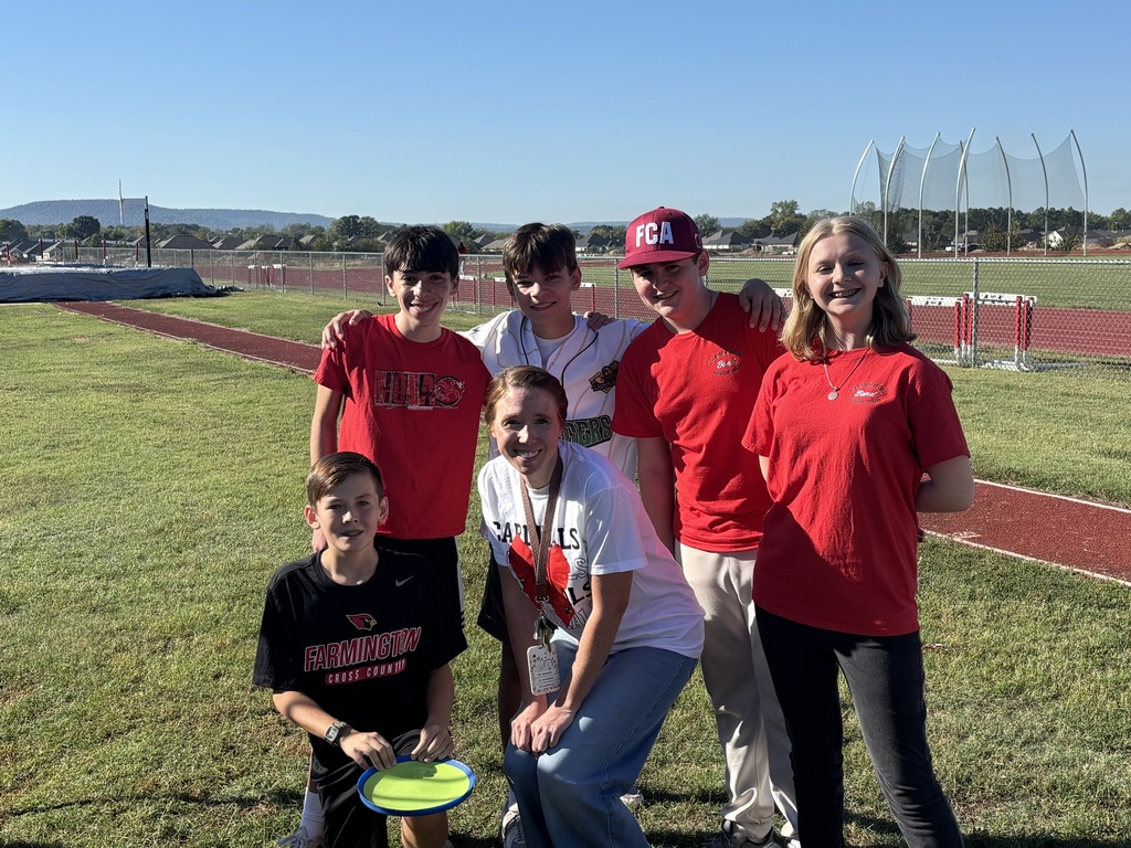 Group of people in red shirts and one in white, standing on a sports field, with one holding a frisbee.